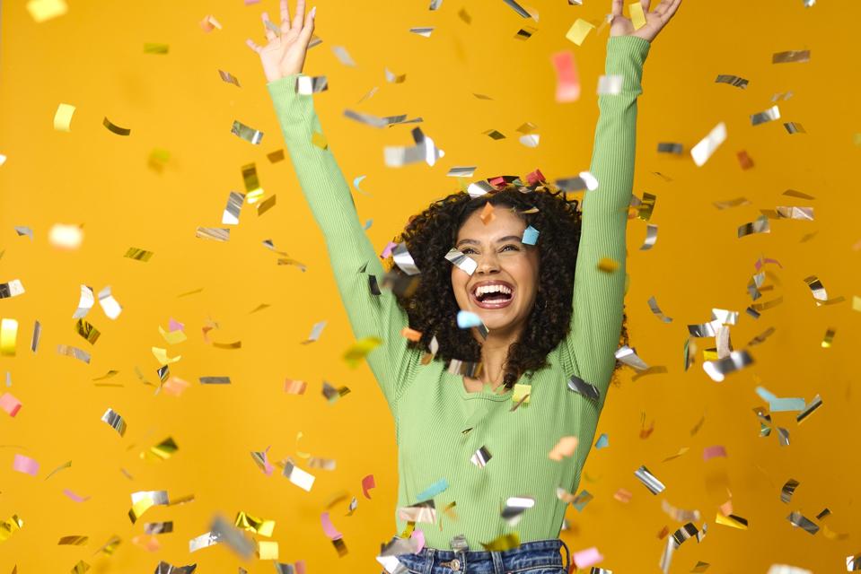 Studio Shot Of Excited Woman Celebrating Big Win Showered In Tinsel Confetti On Yellow Background